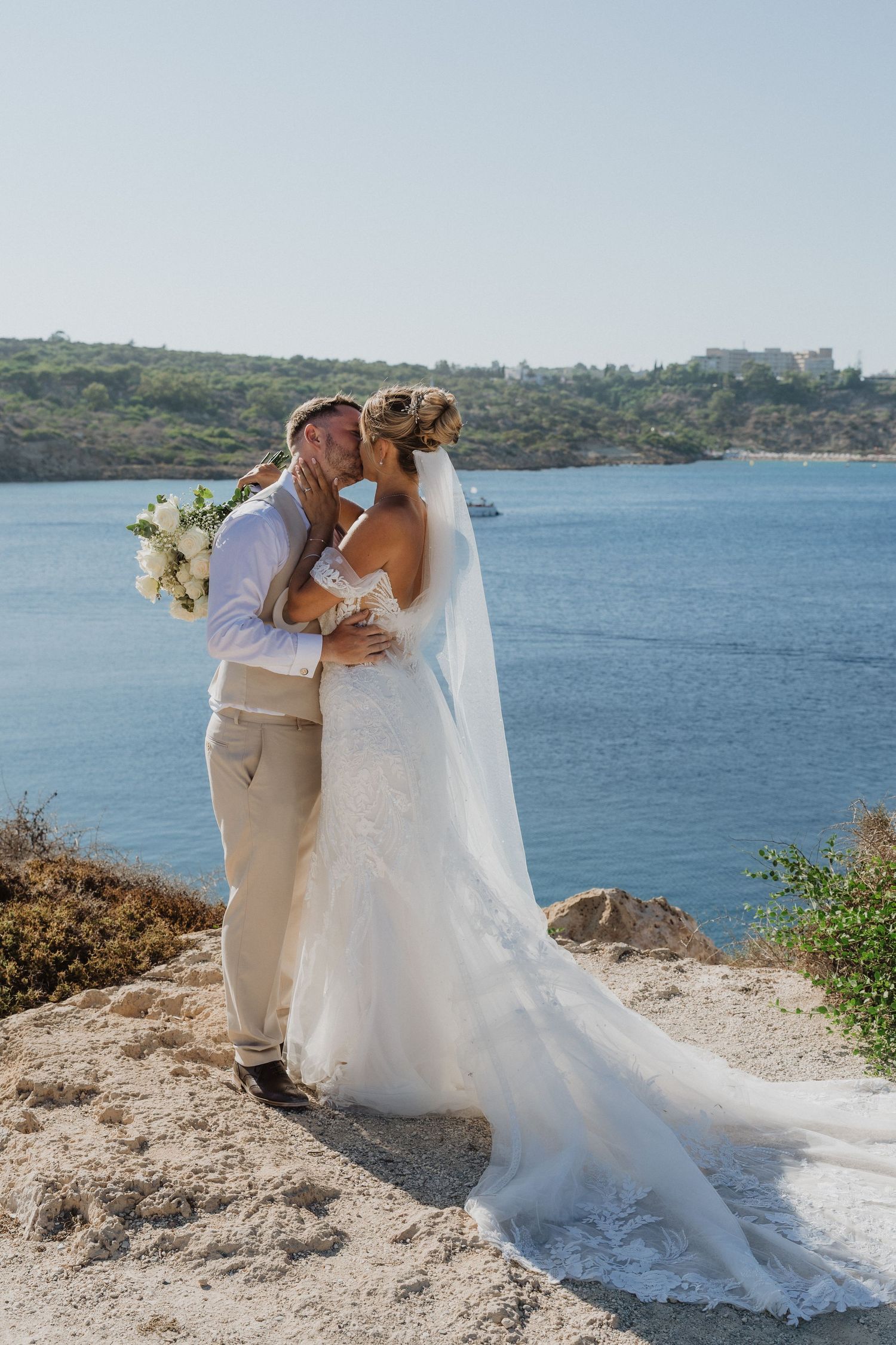 Newlyweds share an intimate moment on a scenic cliff overlooking a beautiful blue ocean bay.