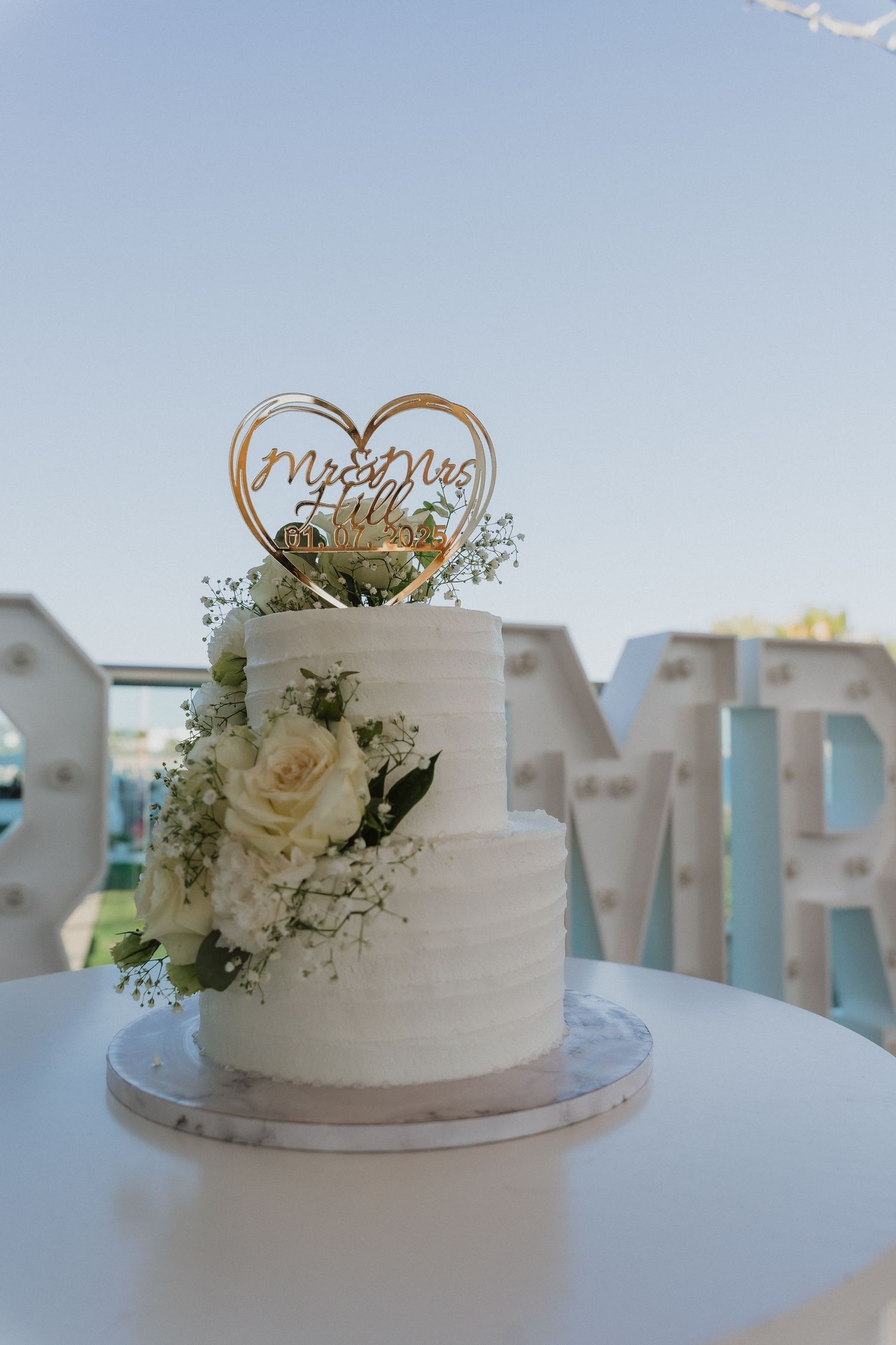 Elegant two-tier white wedding cake decorated with fresh roses and baby's breath, topped with a wooden heart cake topper.