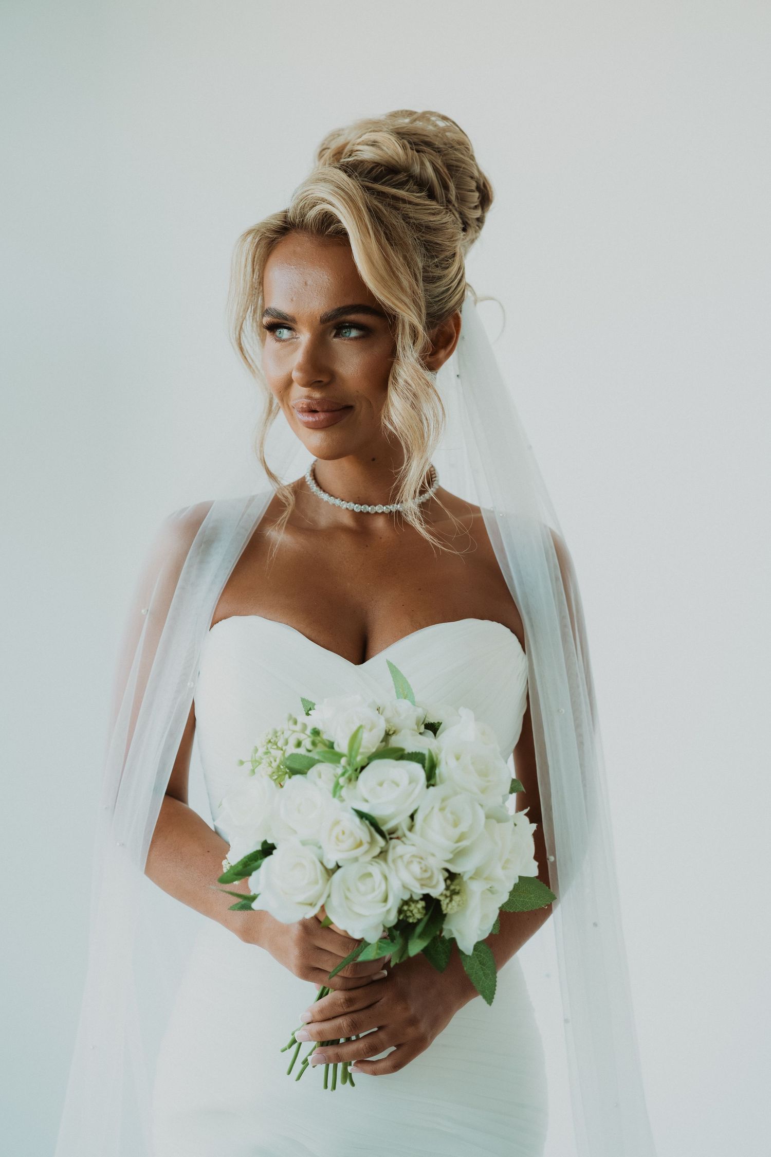 Elegant bride in strapless white gown holding bouquet of white roses, wearing veil and diamond necklace.