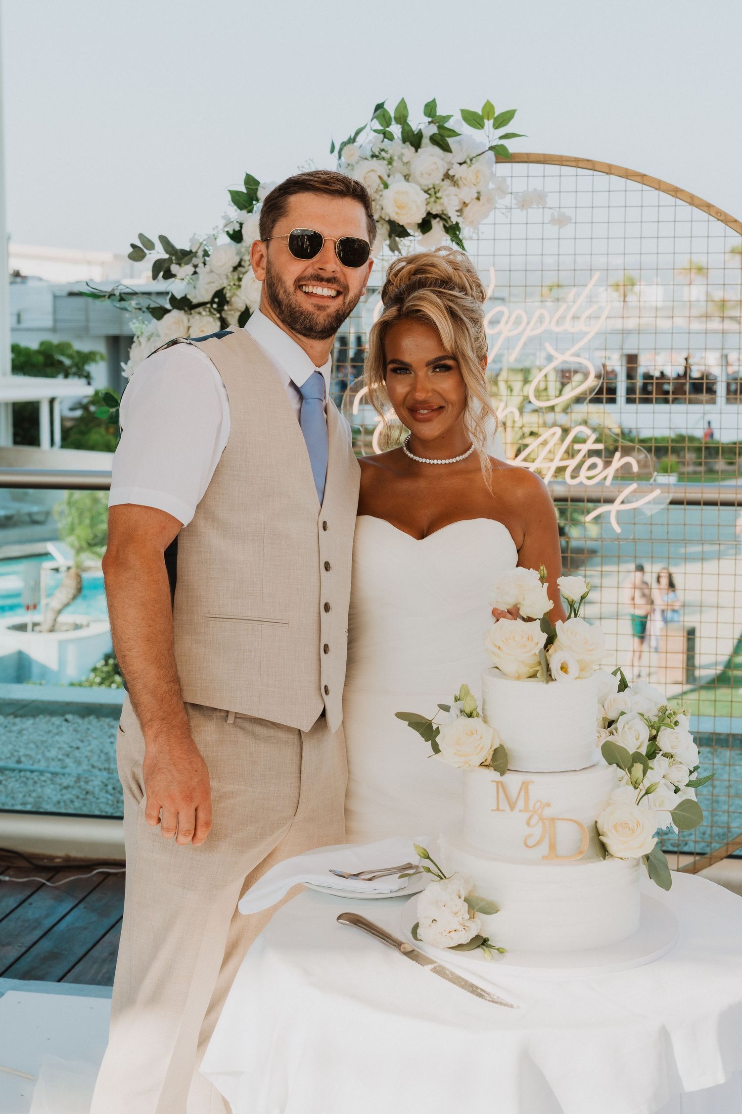 Smiling bride and groom posing beside a white floral wedding cake outdoors at a waterfront venue.