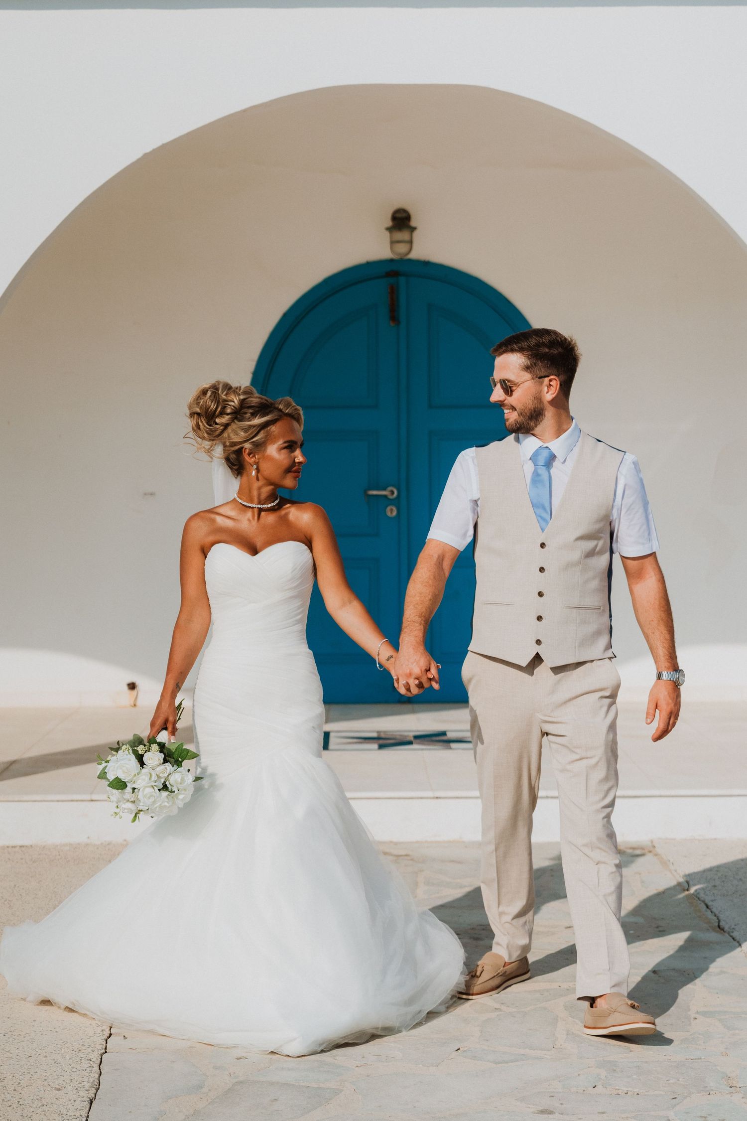 Bride and groom holding hands in front of a blue door and white arch in a Greek island wedding setting.