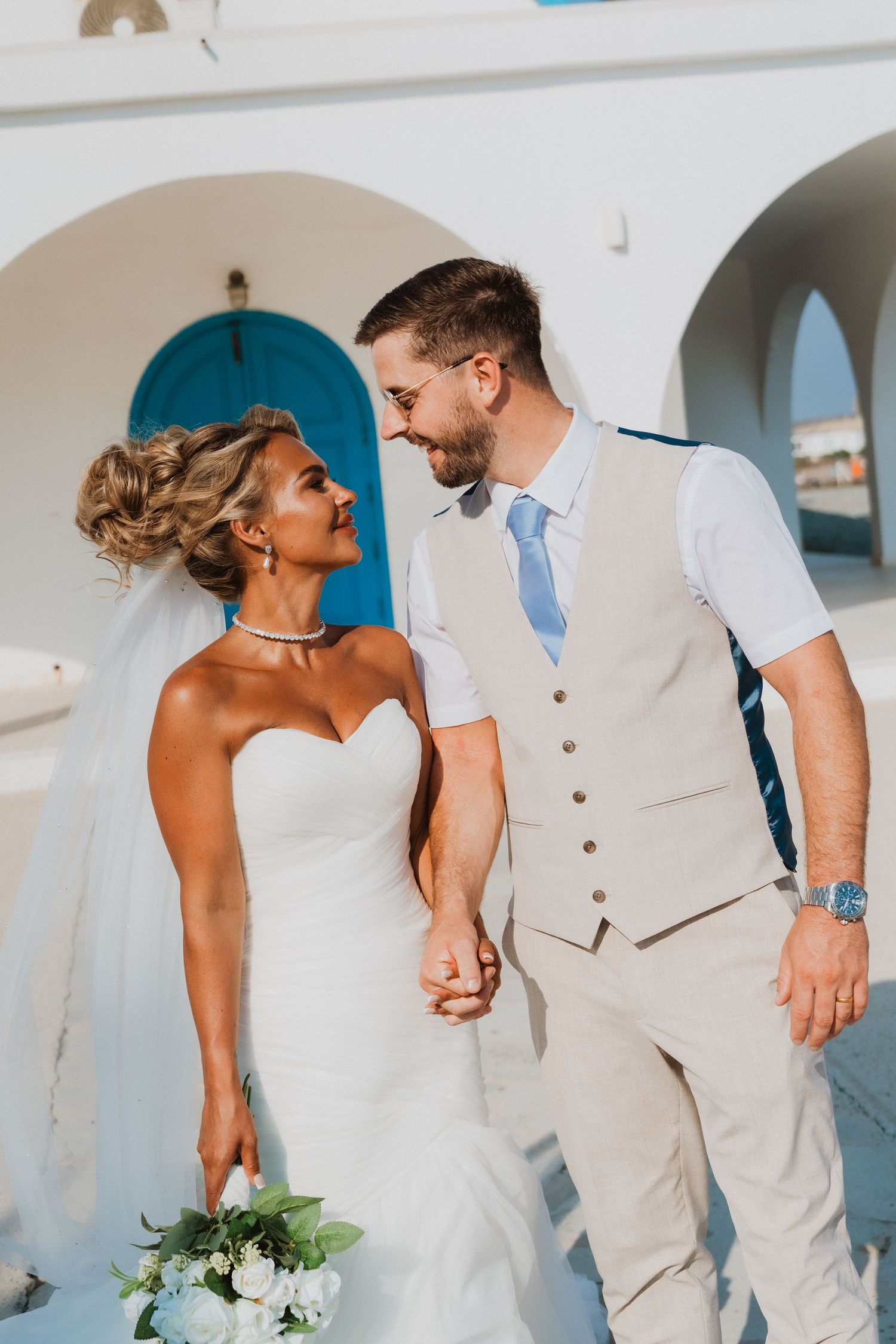 Bride and groom holding hands smiling at each other outside a white Santorini chapel with blue door.