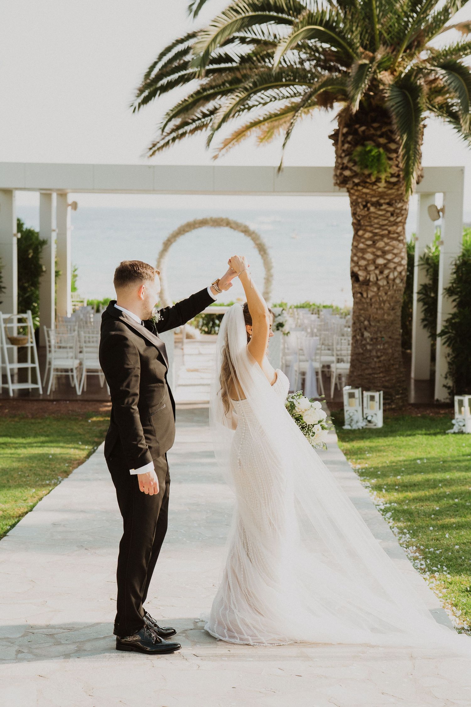 Groom twirls bride in elegant white gown on wedding aisle with palm tree and floral arch backdrop.