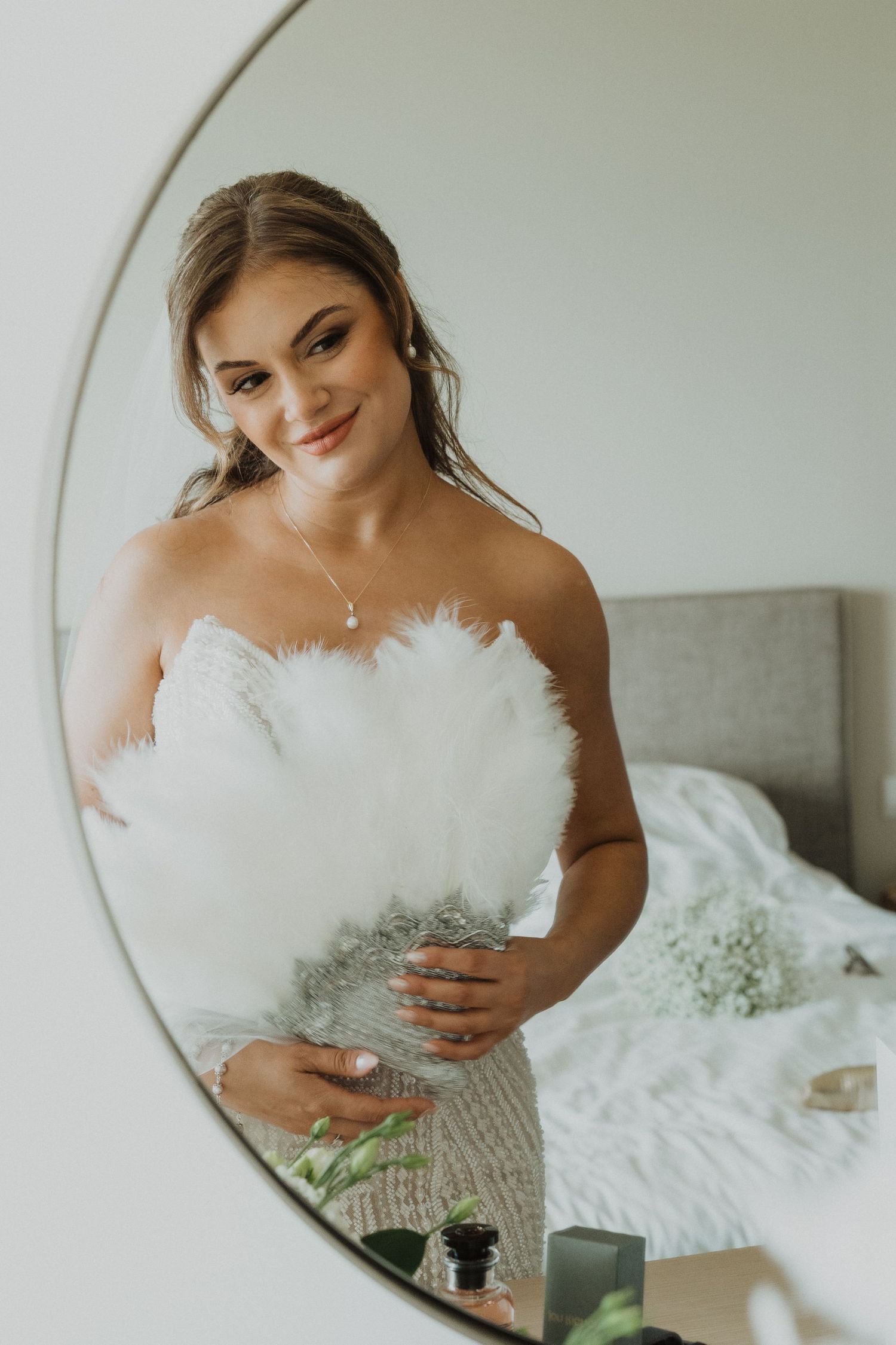 Smiling bride in strapless beaded gown holds white feather bouquet reflected in round bedroom mirror.