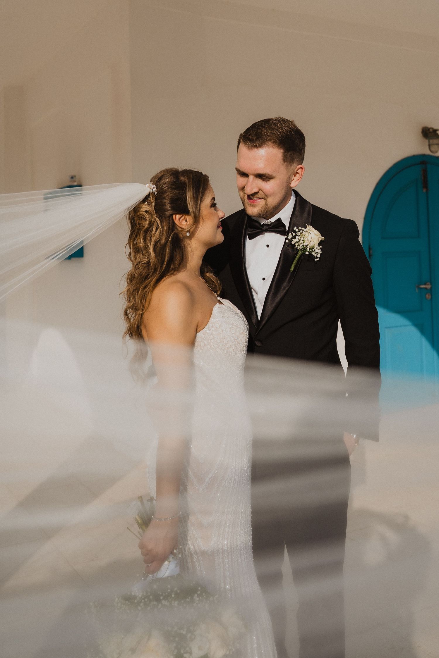 Bride and groom gazing at each other near turquoise door, veil dramatically sweeping across foreground.