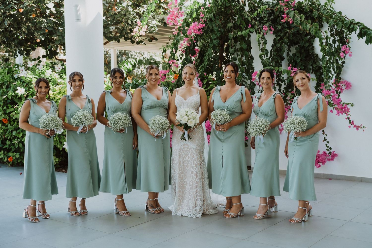 Bridesmaids in sage green dresses pose together at an outdoor wedding venue with pink flowers in the background.