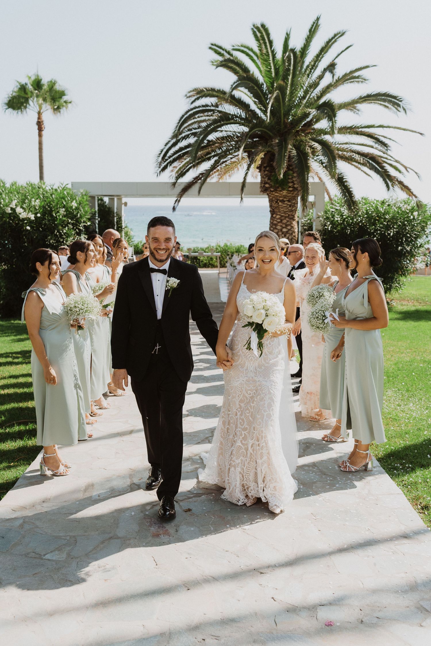 Wedding processional on a white stone walkway with palm trees and ocean views in the background.