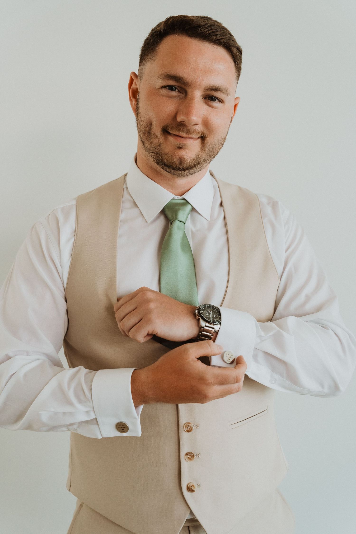 A person wearing a beige vest and mint green tie adjusts their cufflink while posing against a white background.
