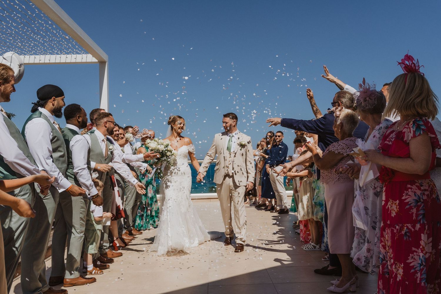 Wedding guests toss flower petals in celebration as newlyweds walk down aisle at beach ceremony.