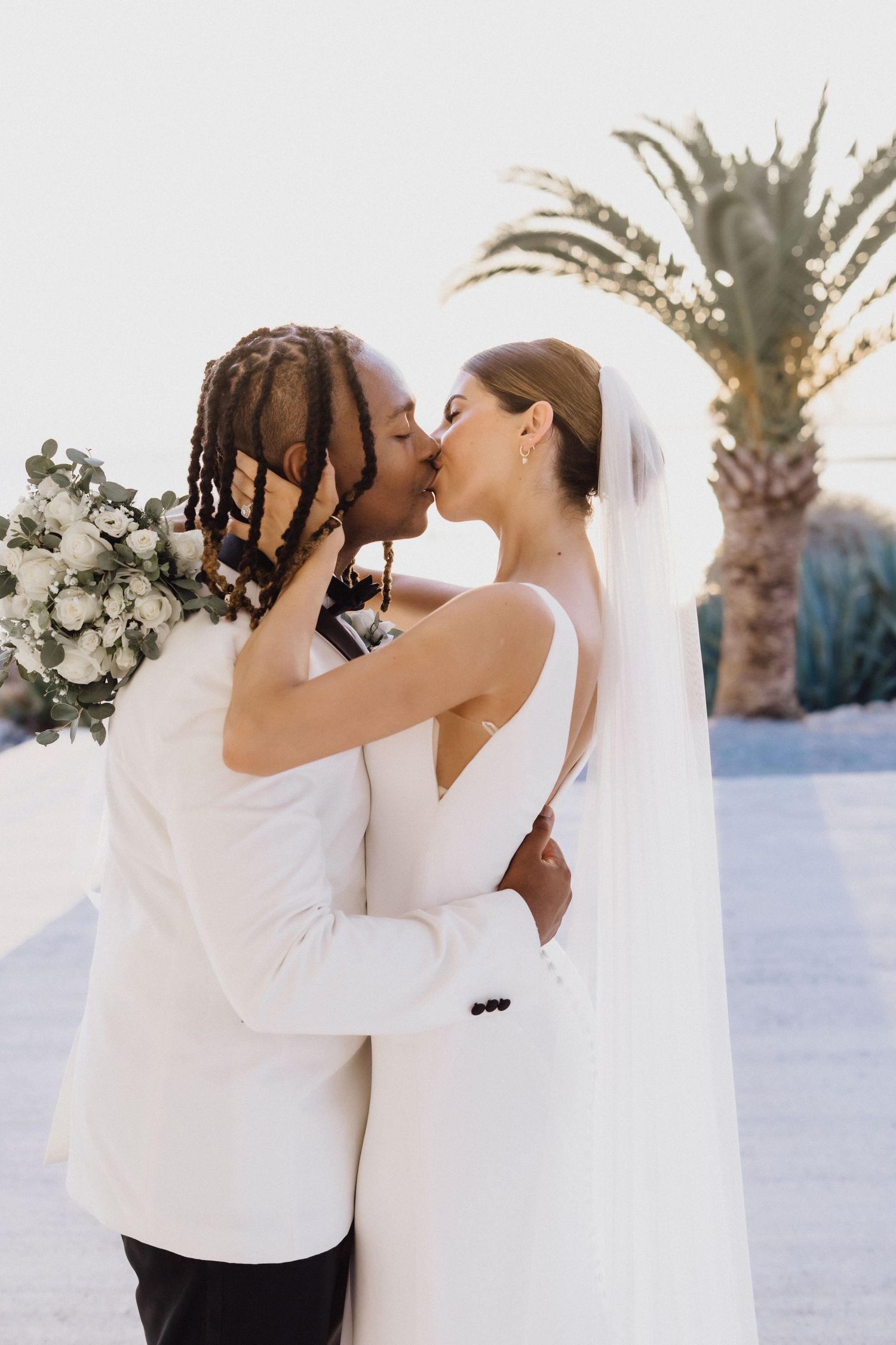 A romantic wedding embrace captured in soft sunlight with palm trees and ocean views.