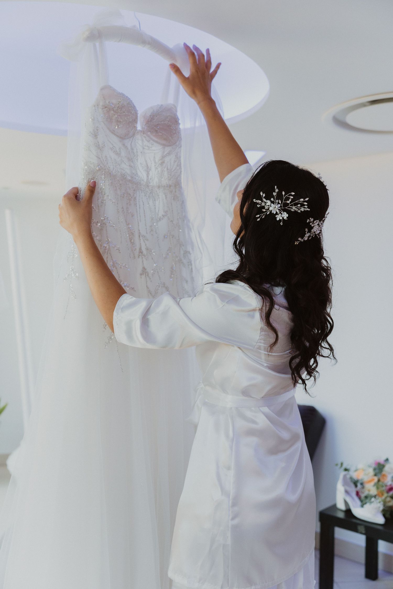 Bride in white robe reaches up to touch her wedding dress, wearing a sparkly hairpiece.