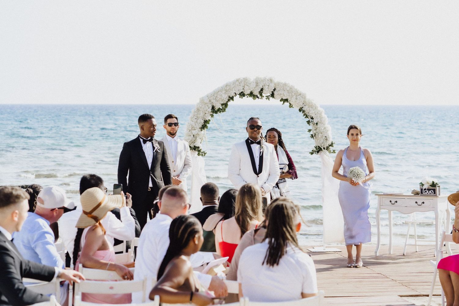A beach wedding ceremony under a floral arch with guests seated on white chairs.