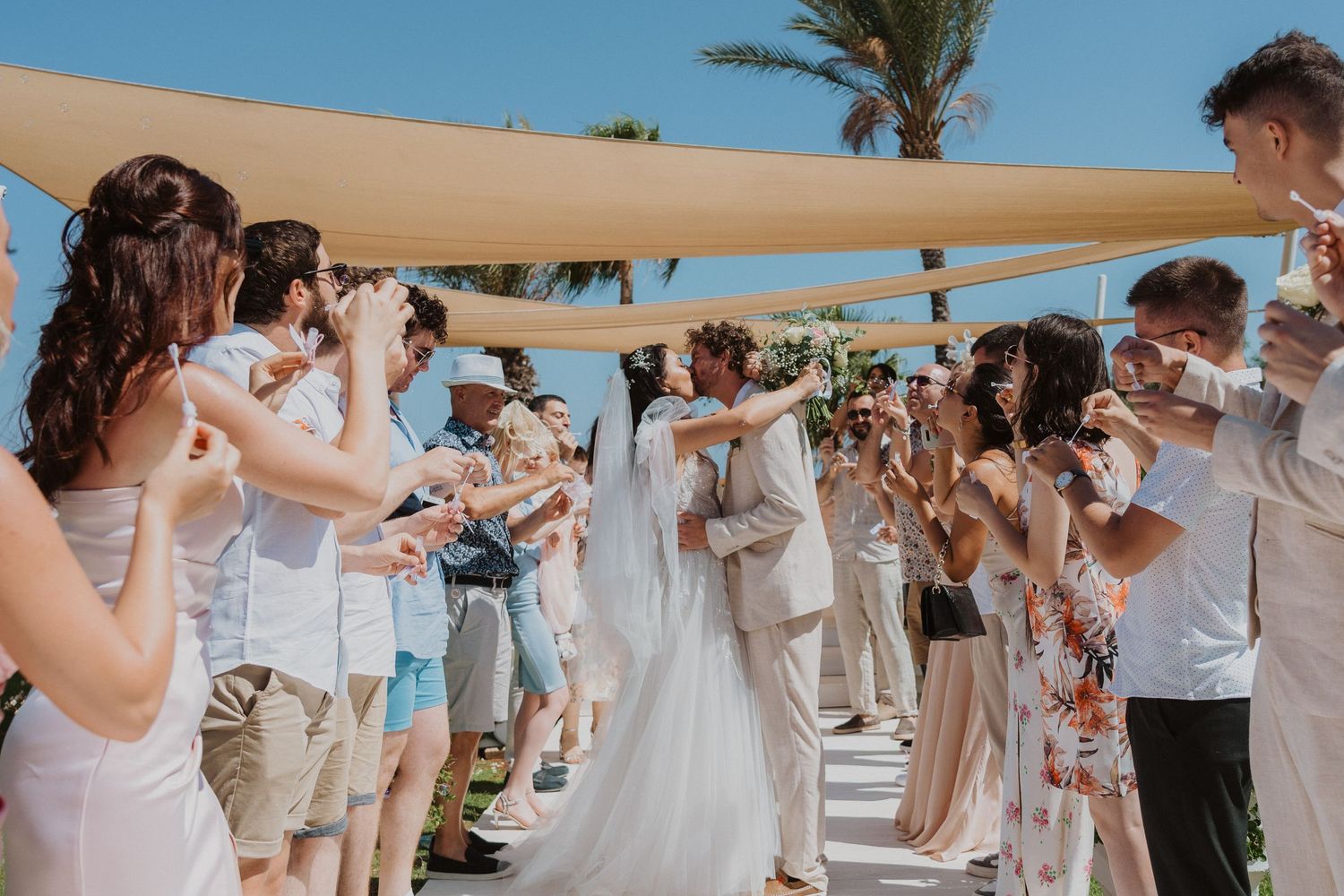 Guests form bubble-filled walkway for newlyweds at beach wedding under tan shade sails.
