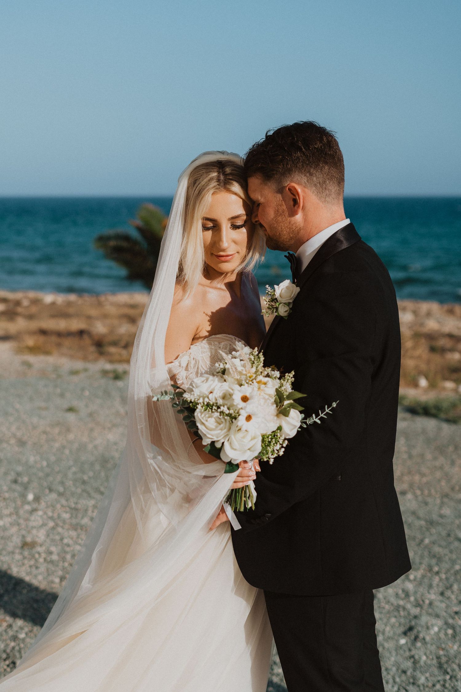 A romantic beachside wedding portrait with white bouquet against turquoise ocean backdrop.