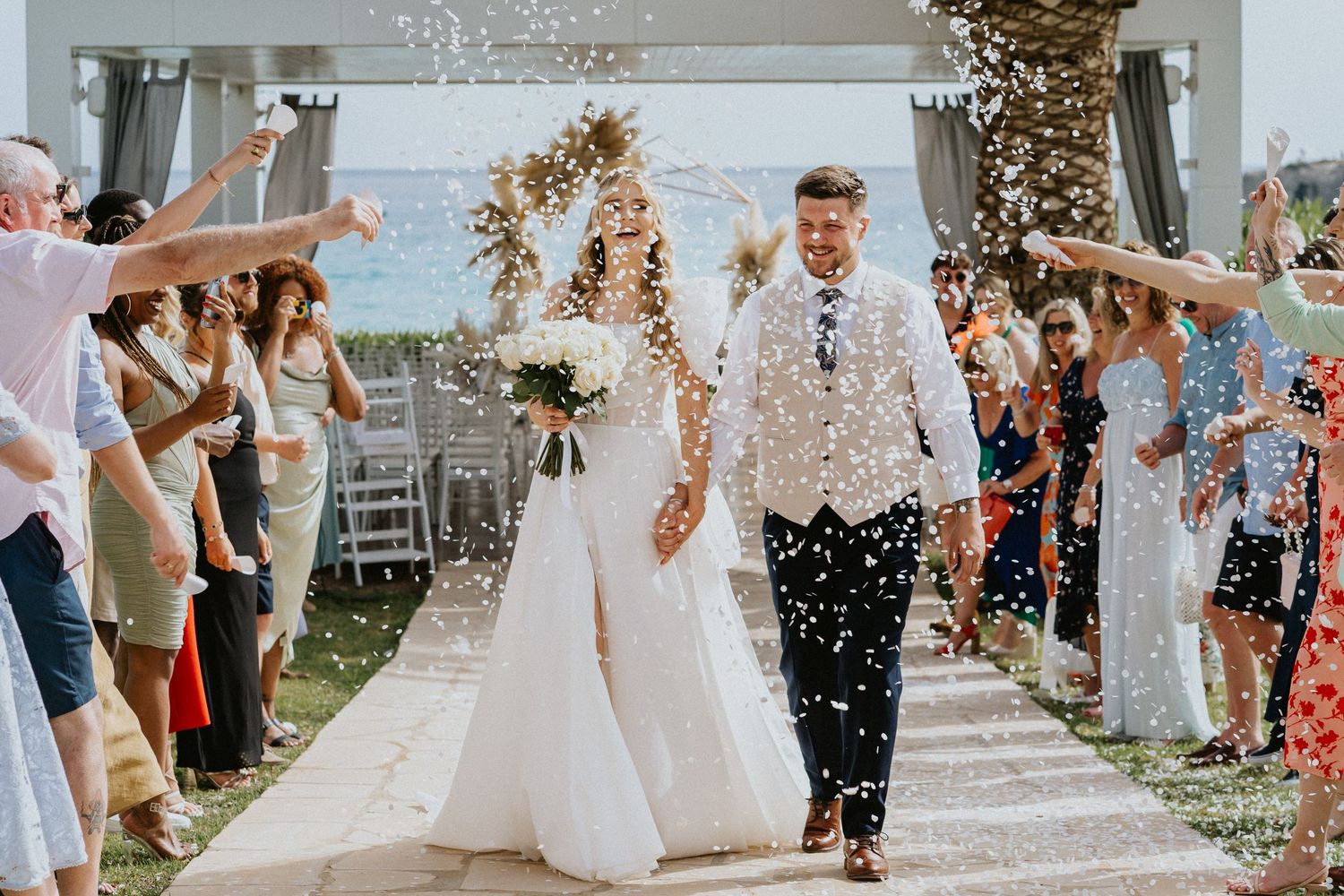 Wedding celebration with guests throwing confetti as newlyweds walk down outdoor aisle with ocean view backdrop.
