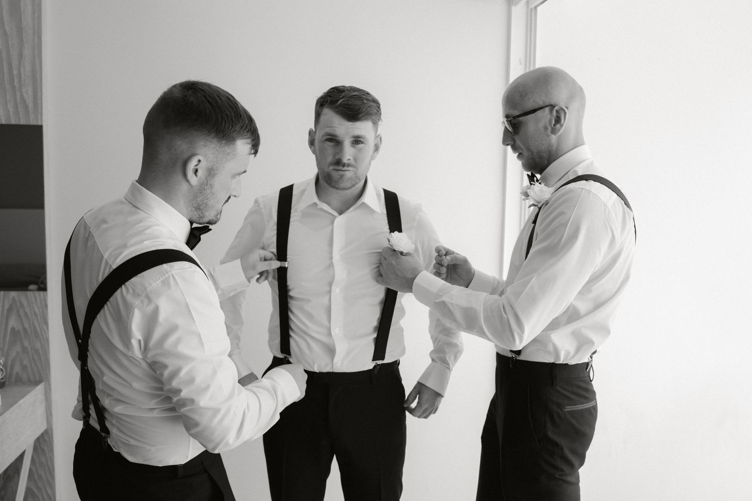 Three men in formal attire with suspenders gather for a black and white candid photo.