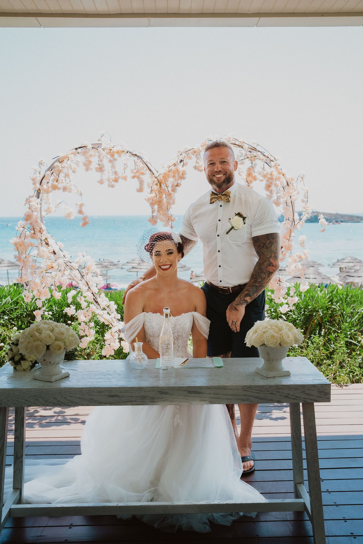 A wedding portrait under a floral arch with ocean backdrop and white flower decorations.
