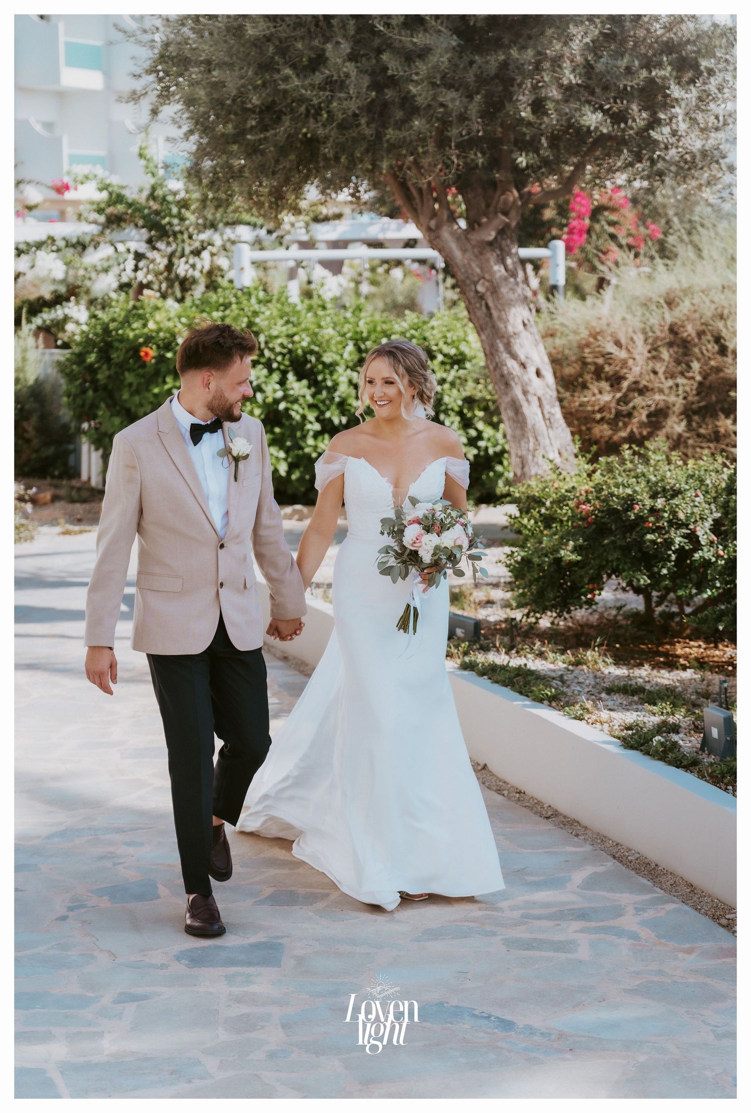 A couple walks hand in hand along a garden path in their wedding attire.