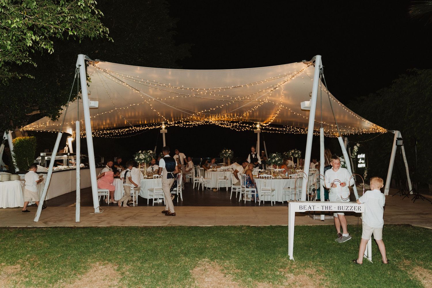 An outdoor wedding reception tent glows with fairy lights as guests dine at tables underneath.