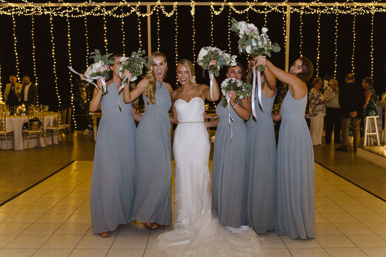 Bride and bridesmaids pose playfully with bouquets under golden string light decorations.