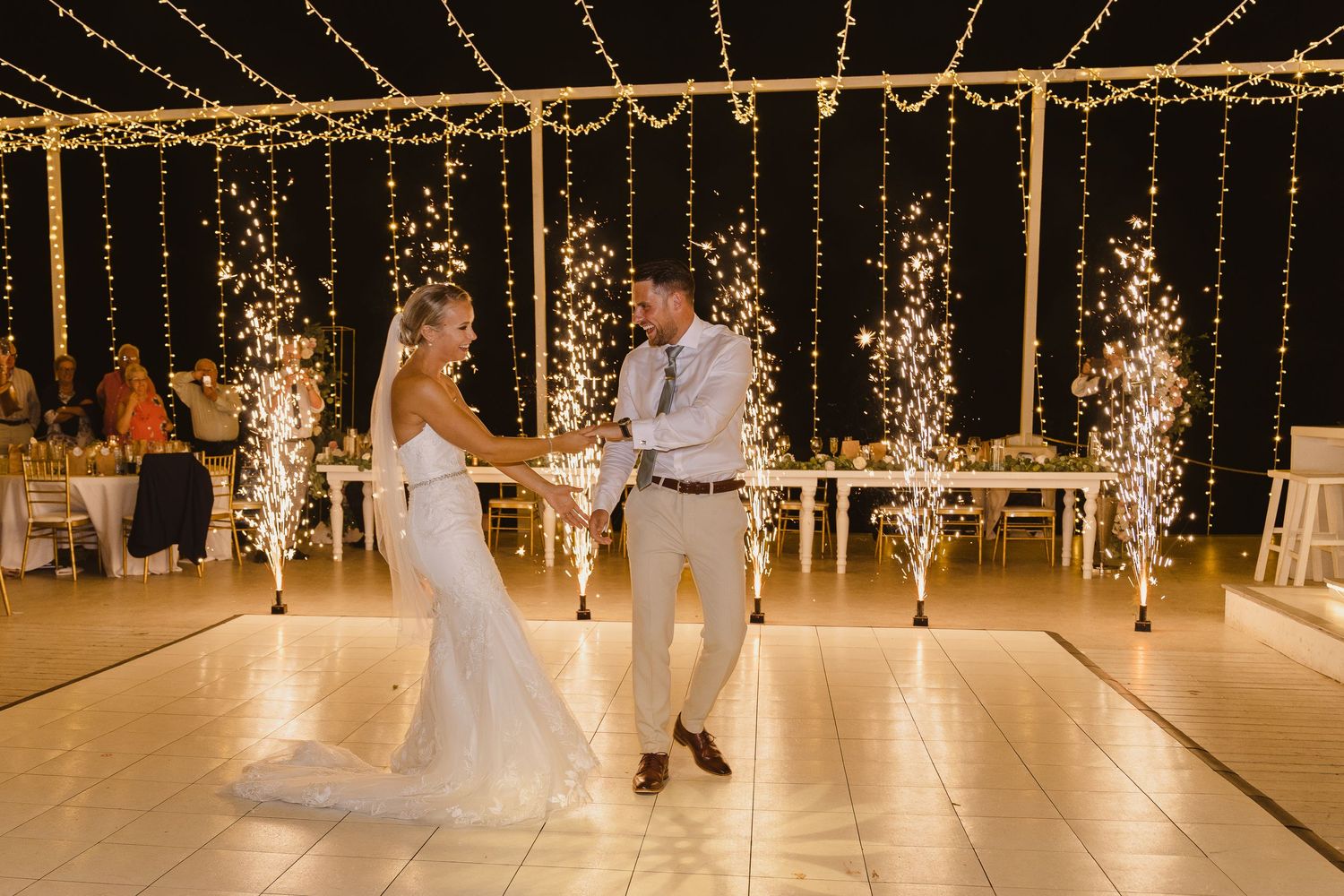 Newlyweds dance on illuminated floor with cascading sparkler effects and fairy lights in background.