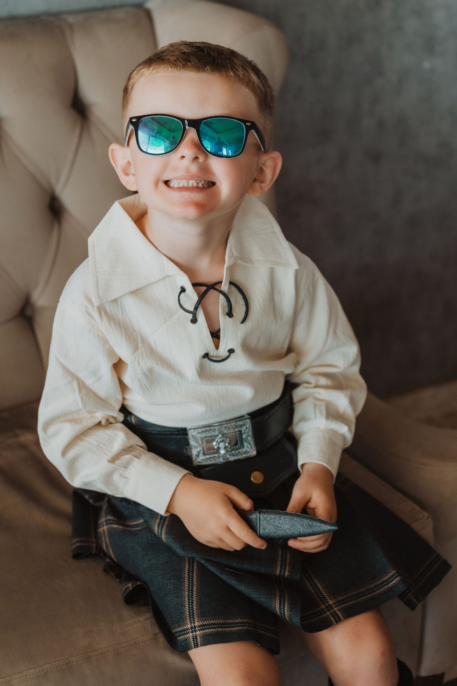 A young boy in Scottish attire and sunglasses sits on a beige couch.