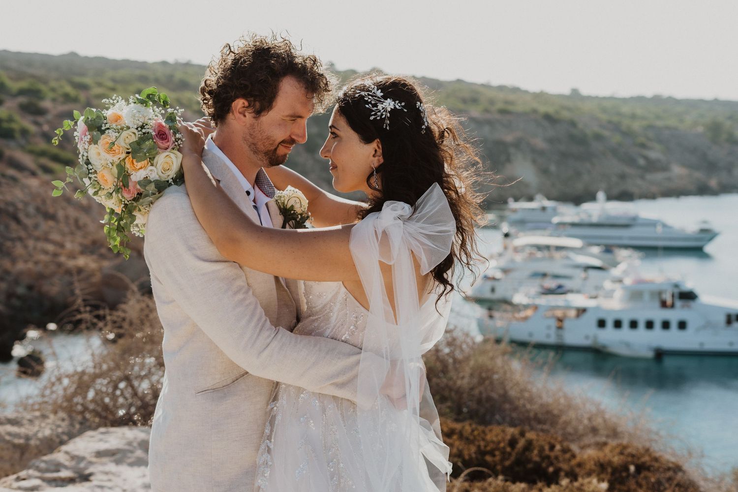 A romantic wedding photo showing a couple dancing on a coastal overlook with boats in the background.