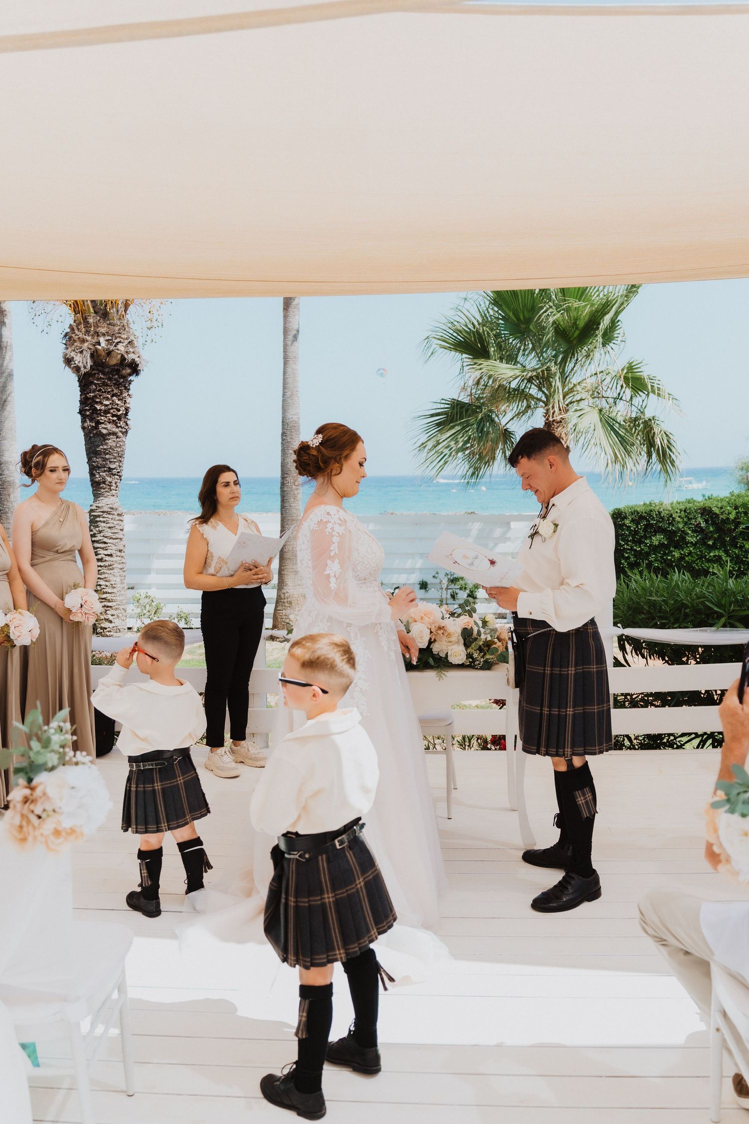 Children in traditional Scottish kilts watch a beachside wedding ceremony under a white canopy with palm trees.