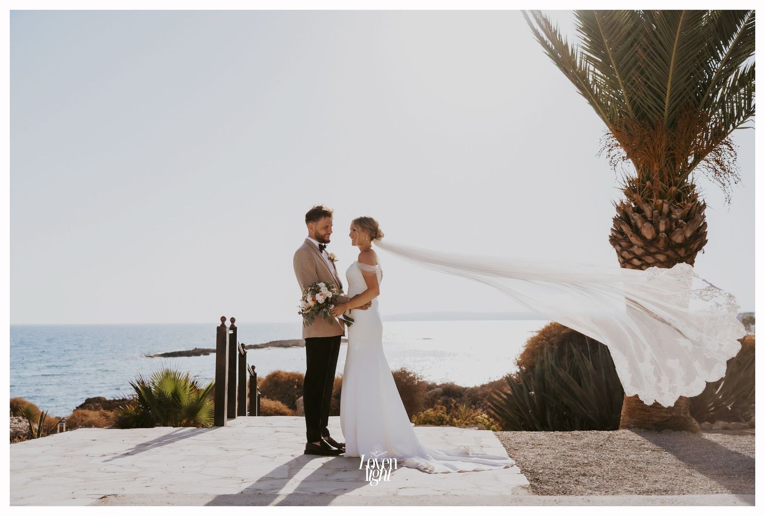 Wedding photo with flowing veil against coastal scenery and palm trees.
