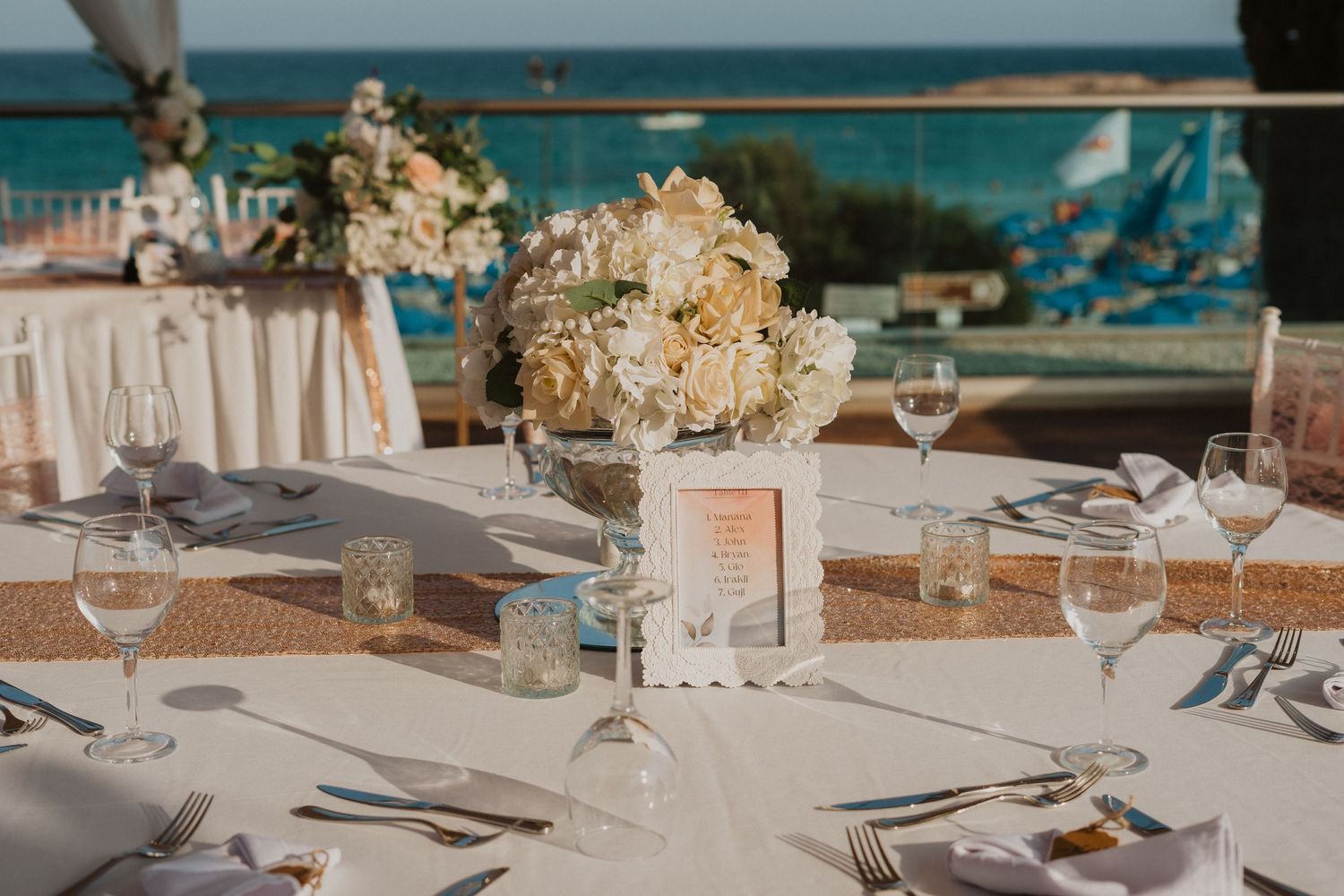 Wedding reception table with white hydrangea centerpiece, gold runner, and ocean view backdrop.