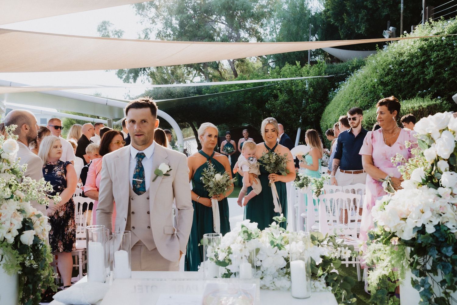 A wedding ceremony setup outdoors with white floral decorations and guests seated under shade sails.