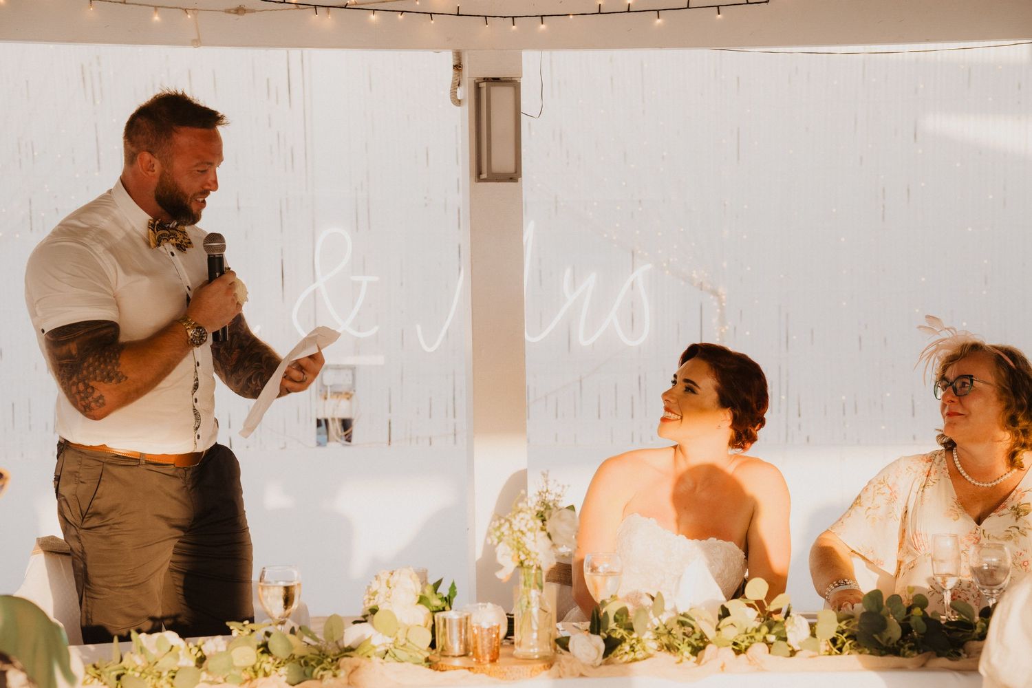 Wedding party at reception with floral centerpieces and string lights overhead.