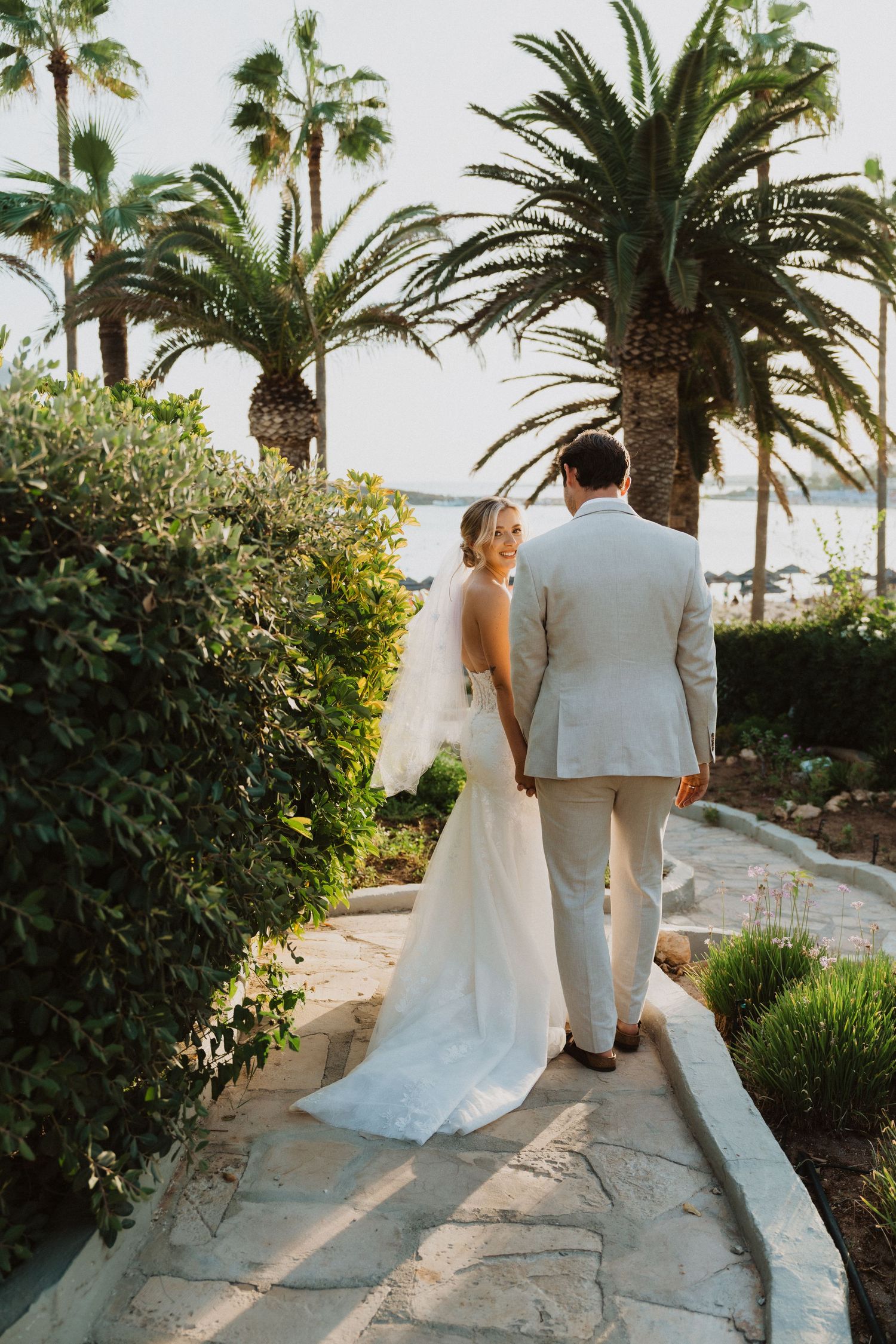 A couple in wedding attire walk along a palm-lined path overlooking the ocean at sunset.