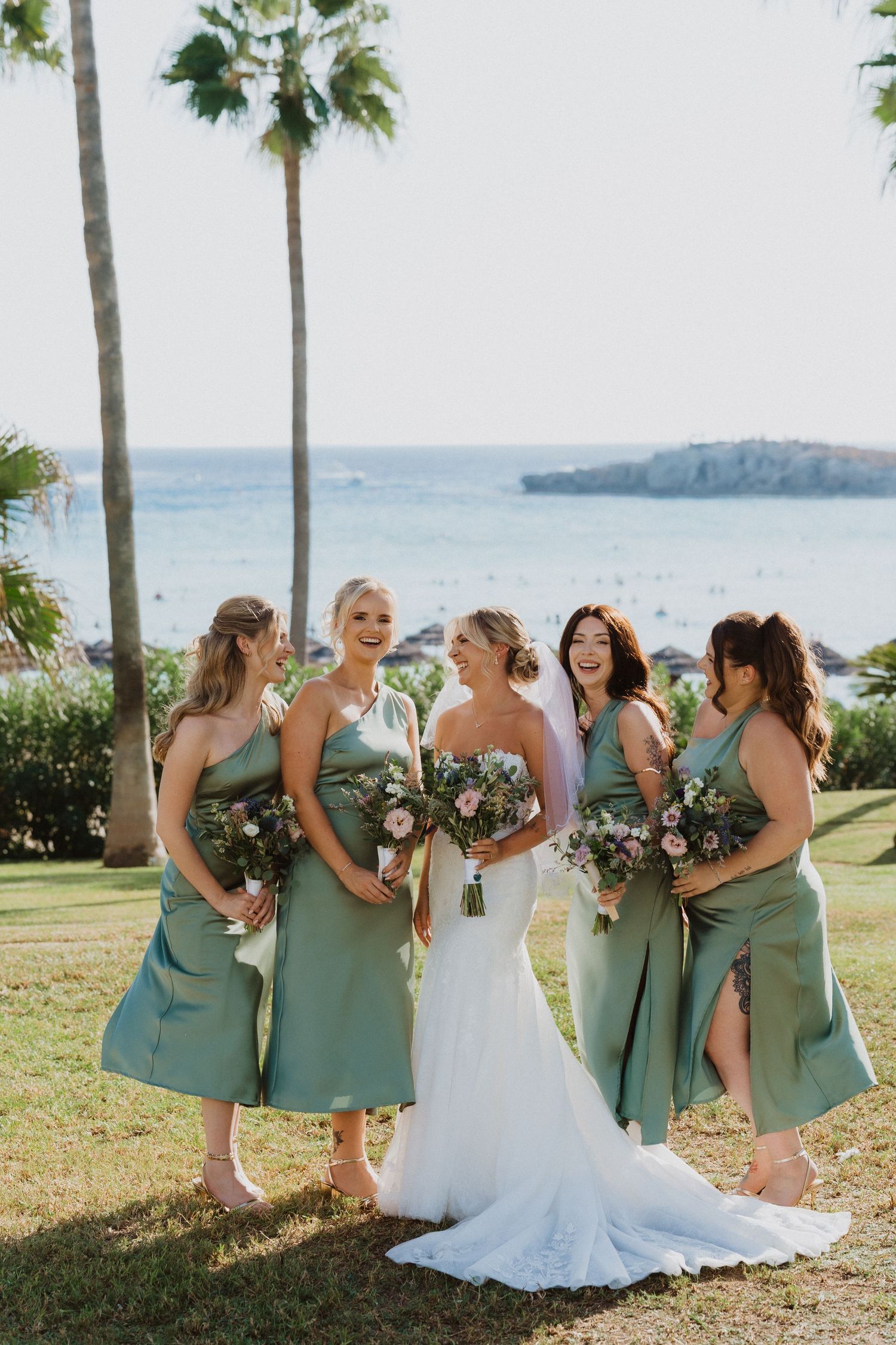 Bridesmaids in seafoam green dresses pose with bride in white gown against tropical oceanside backdrop.