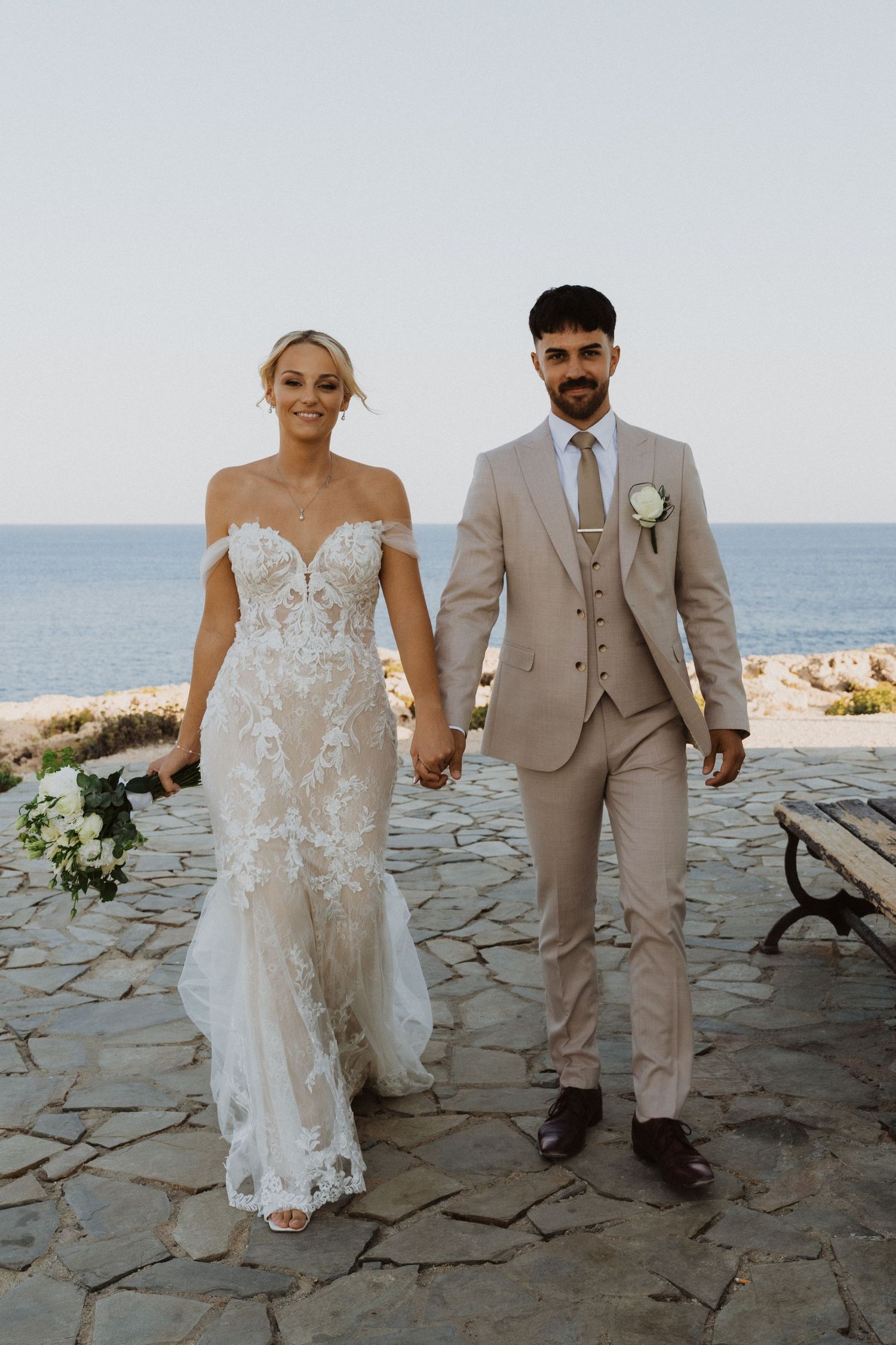 Couple walks hand in hand on stone terrace by Mediterranean seaside.