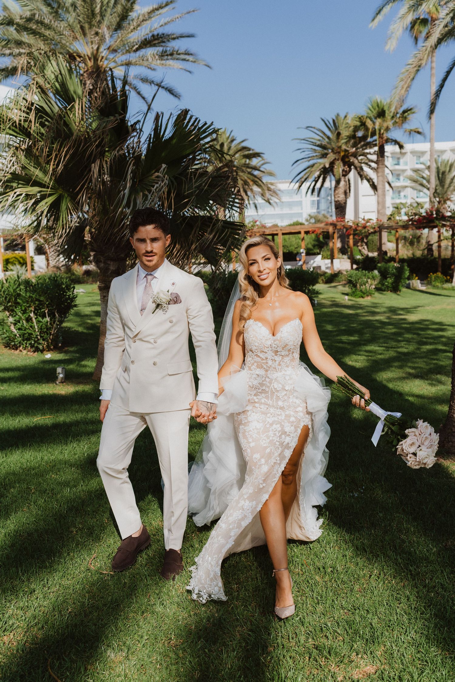 Couple in white attire walking together on a palm-lined lawn during a tropical destination wedding celebration.