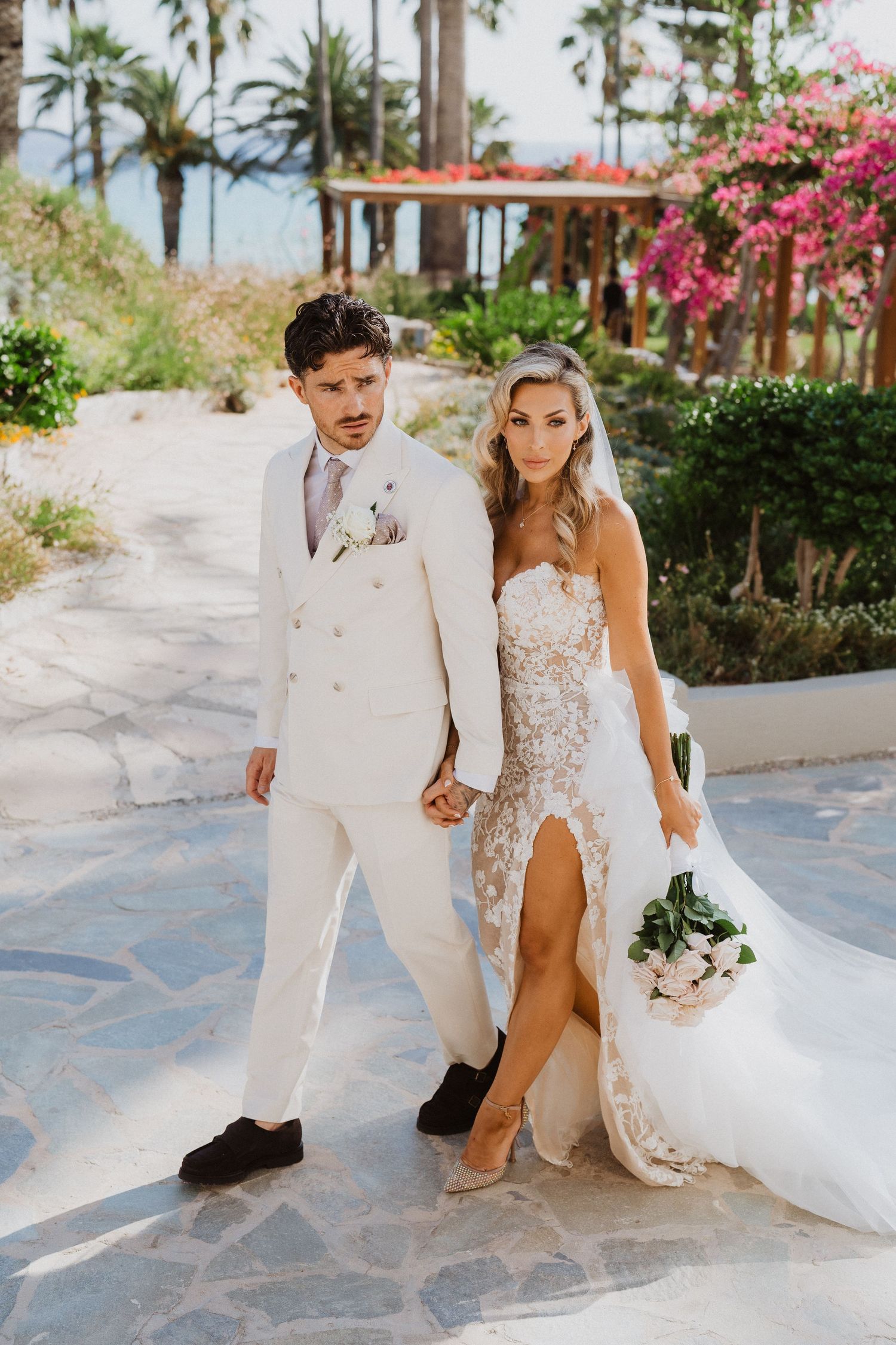 Couple in white attire walking along a stone pathway surrounded by tropical palm trees and pink bougainvillea flowers.