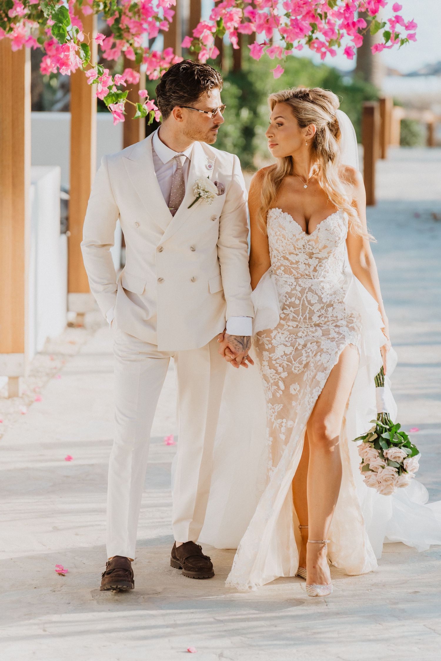 Couple in elegant white wedding attire walking together on a paved pathway with pink bougainvillea flowers overhead.