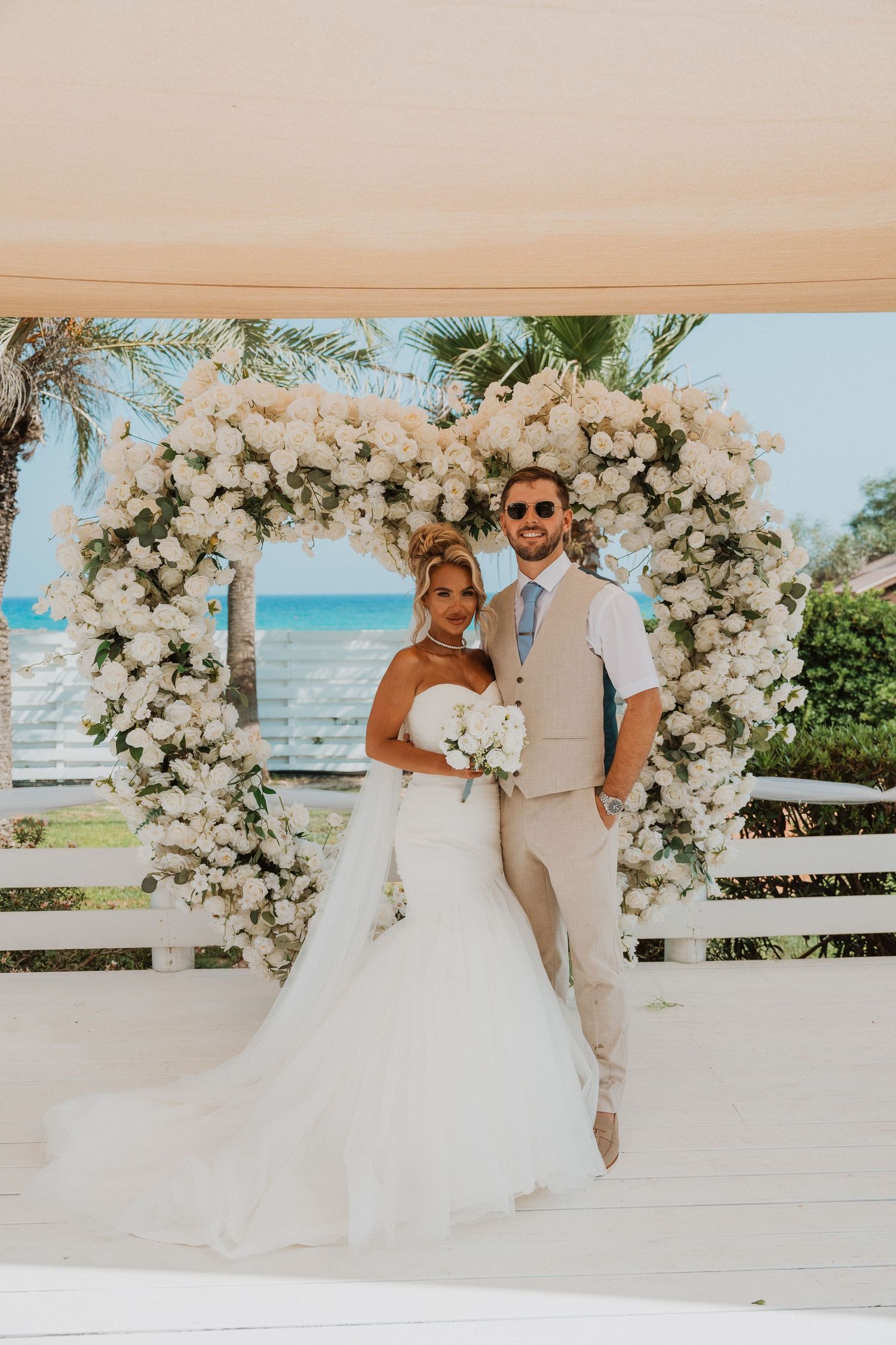 Bride and groom posing under a white floral arch at a beachside wedding ceremony.