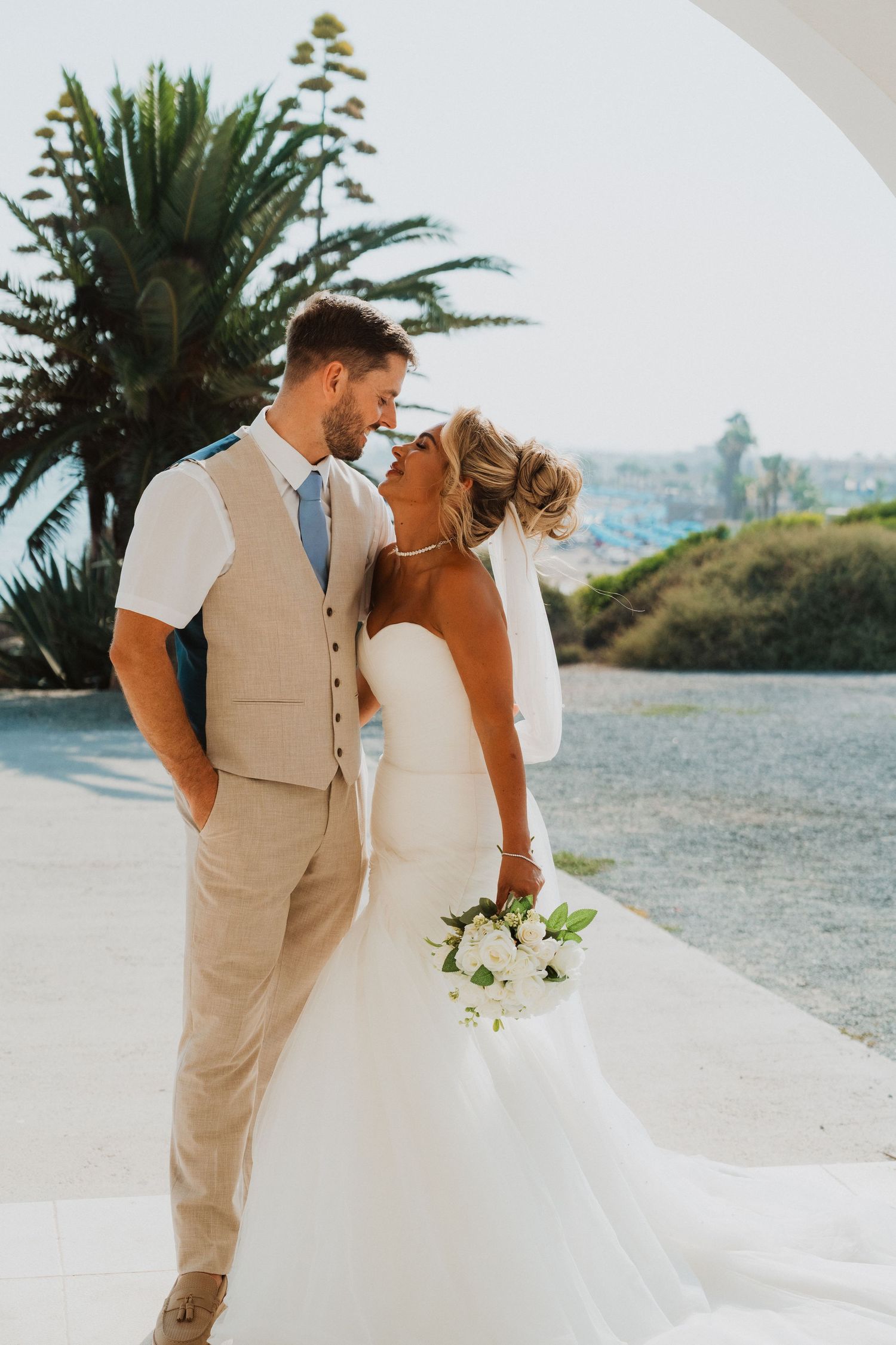Bride and groom share a romantic moment outdoors, she holds white bouquet, he wears tan suit, coastal backdrop.