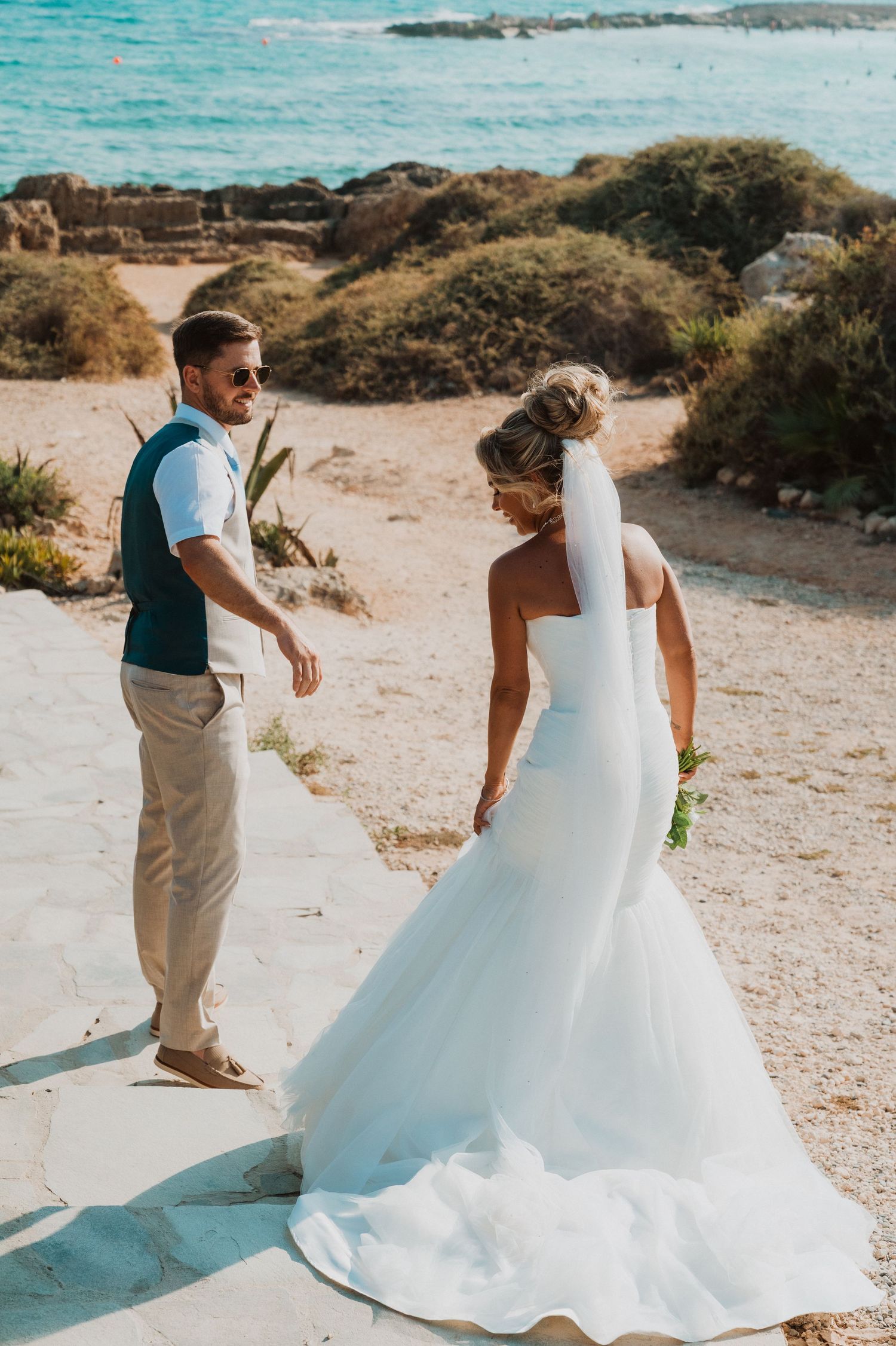 Bride in white mermaid gown and groom in teal vest share a moment by the sea on their wedding day.