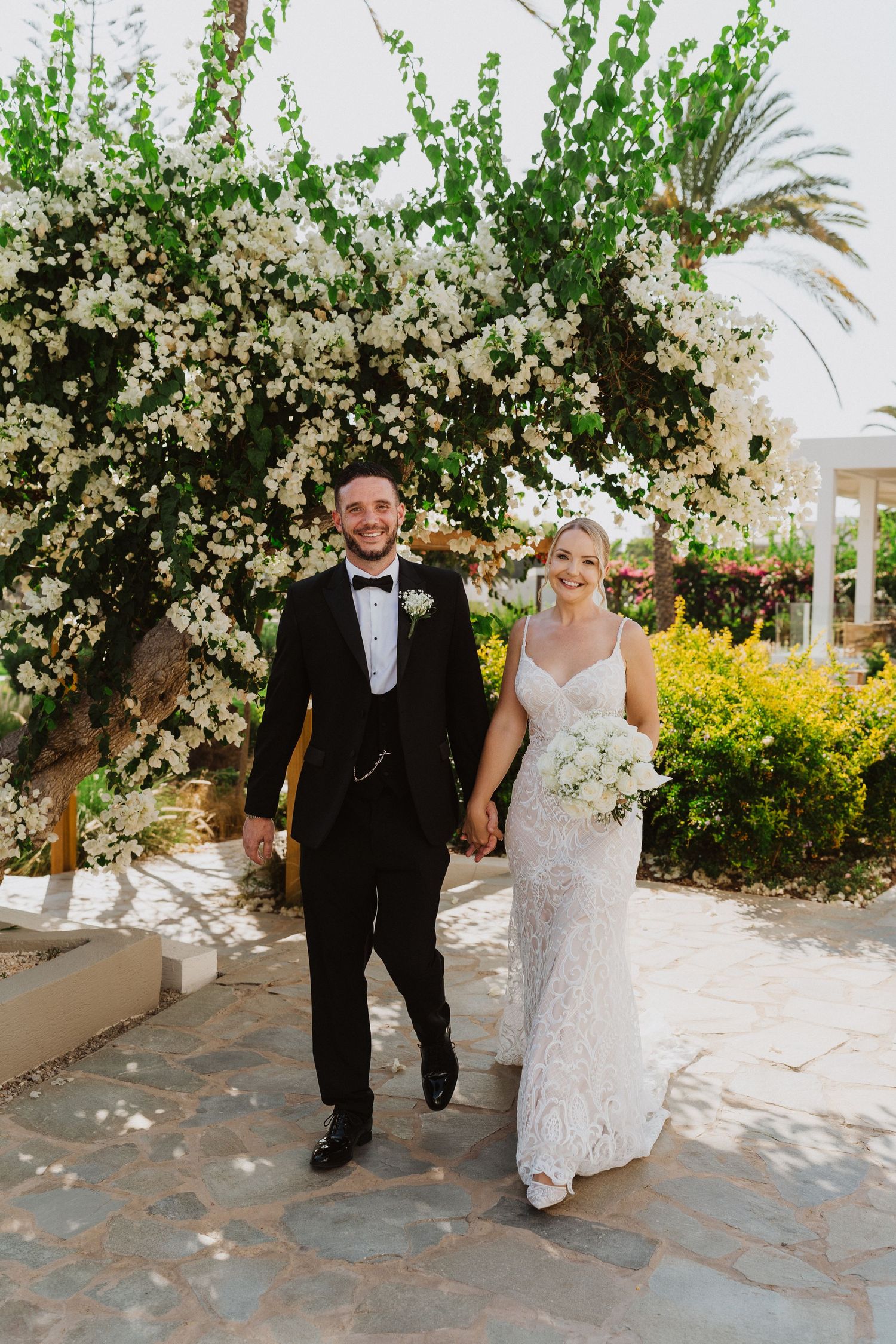 A couple walks together through a garden path with blooming white flowers and palm trees.