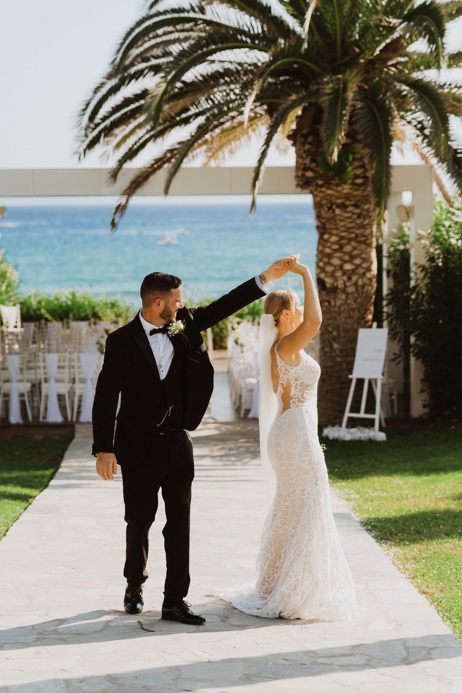 A couple in wedding attire dance together on a palm tree-lined beachfront terrace with ocean views in the background.