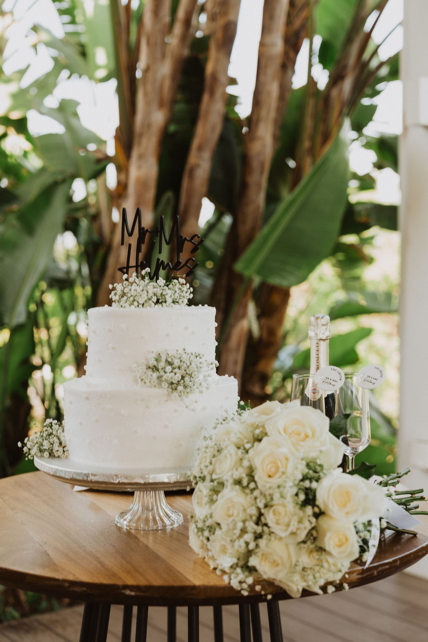 Elegant white wedding cake with floral decorations displayed on a wooden table with tropical foliage in the background.