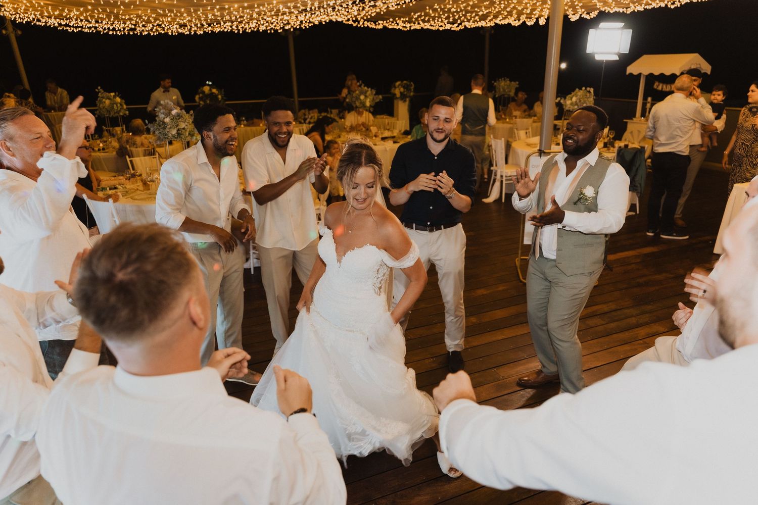 Wedding guests dance together on the dance floor at an evening reception with warm golden lighting.