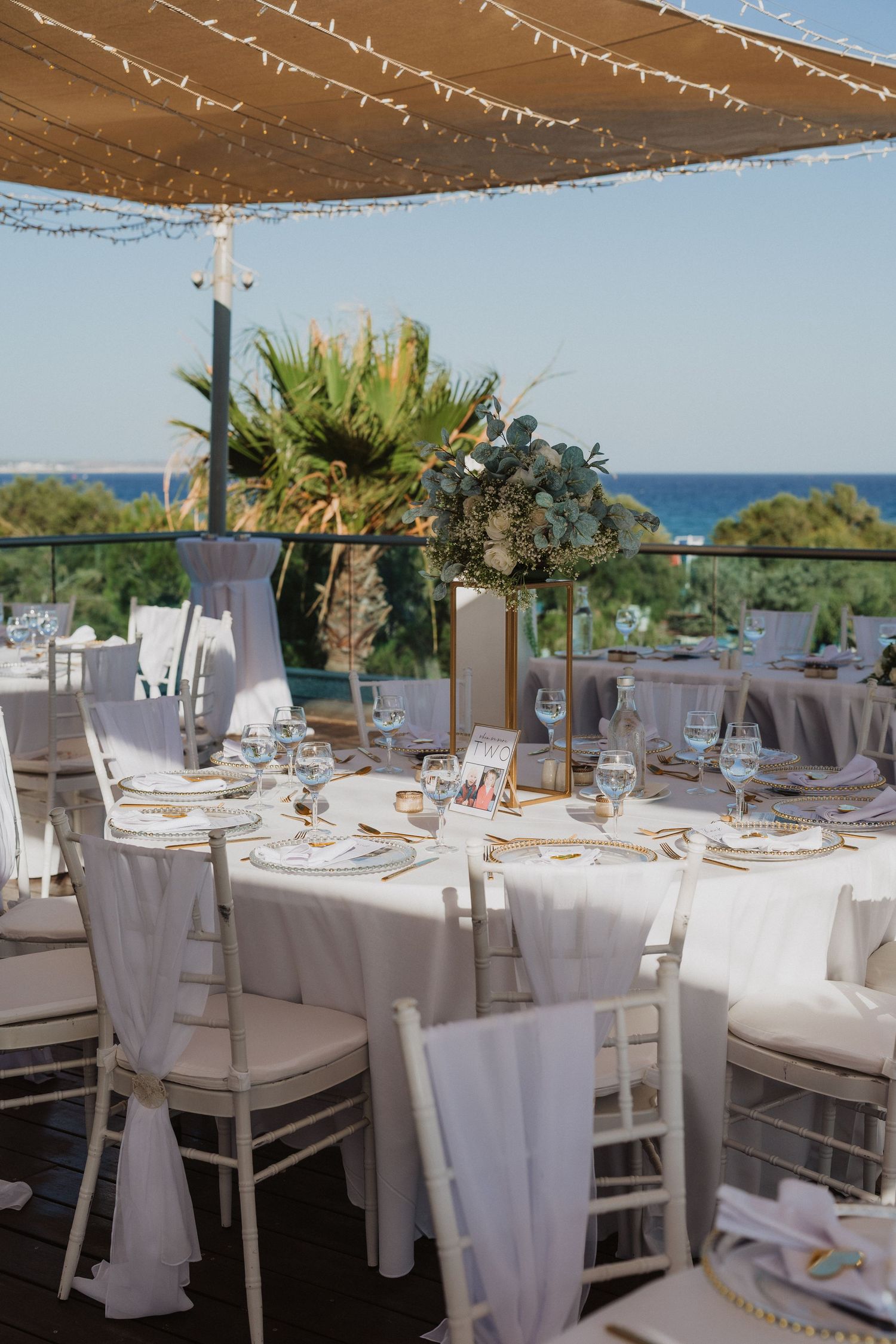 Elegant outdoor wedding reception setup with white-clothed tables and chairs overlooking the ocean under a covered terrace.