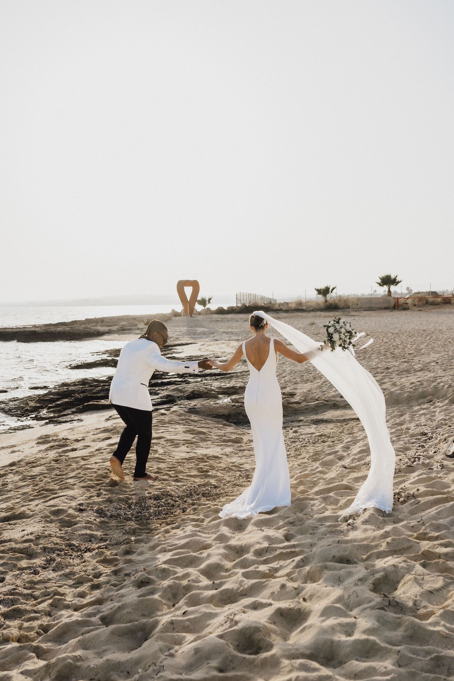 Wedding couple walking on sandy beach with veil flowing in the wind.