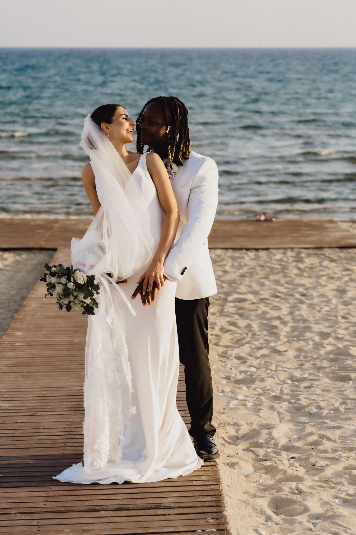 Newlyweds share intimate moment on wooden boardwalk with ocean backdrop.