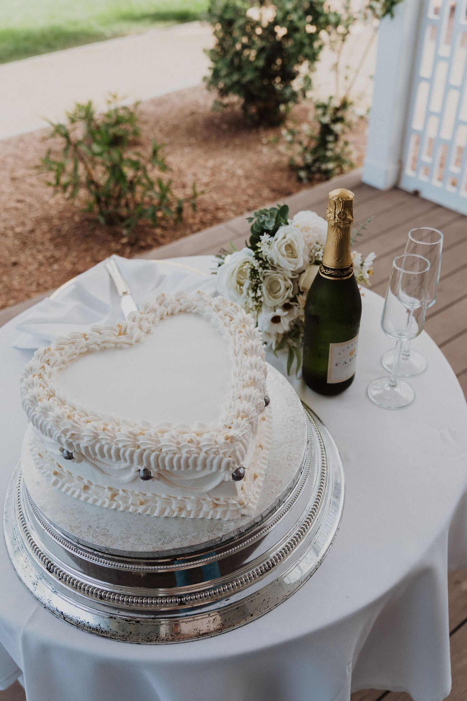 Heart-shaped white wedding cake with champagne bottle and glasses on round table.