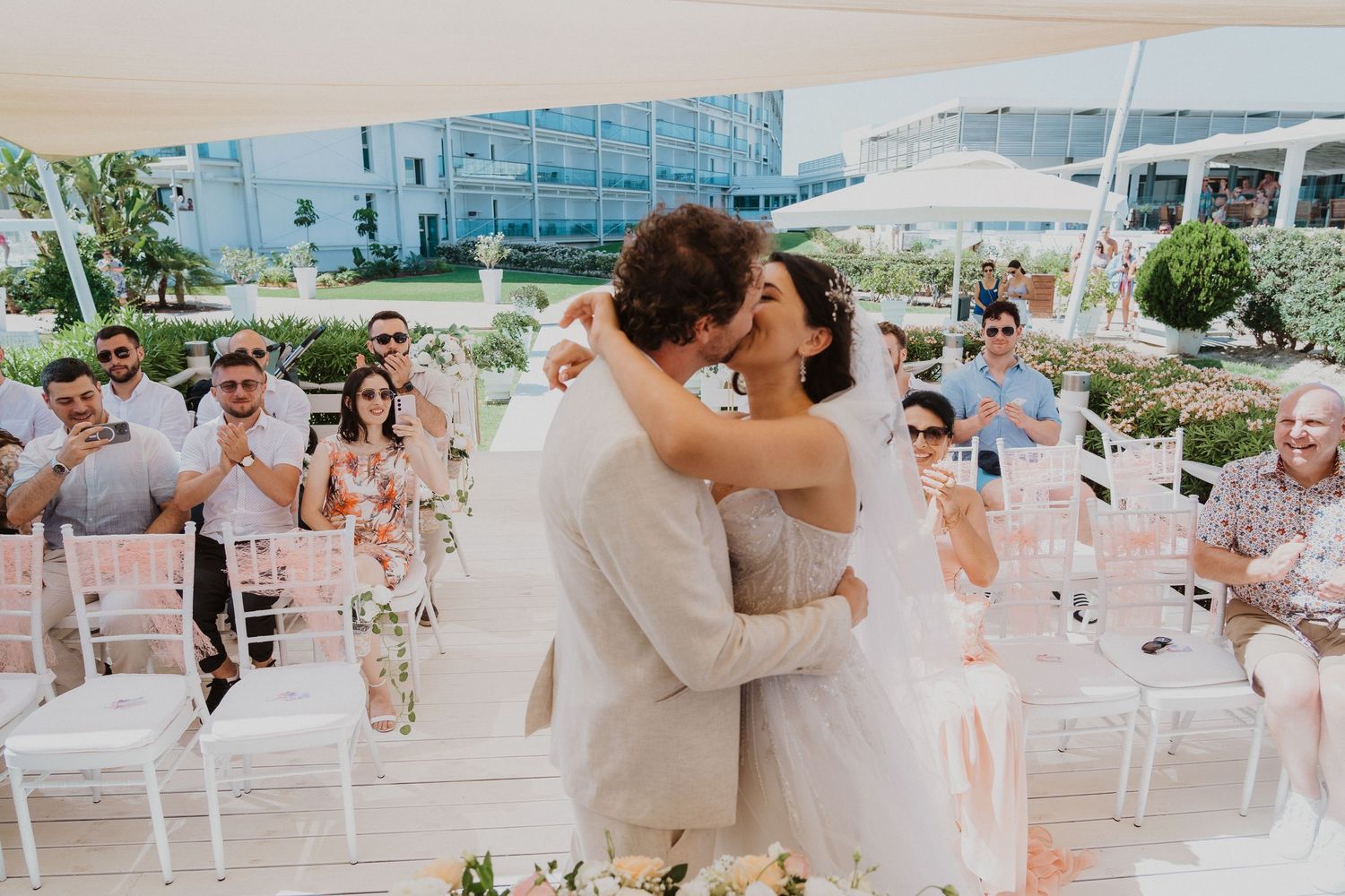 First kiss as newlyweds during outdoor ceremony while guests applaud.